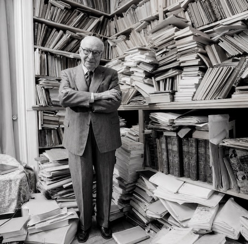 A photograph of Dumézil in a suit, in front of a chaotic mess of books on shelves and the floor
